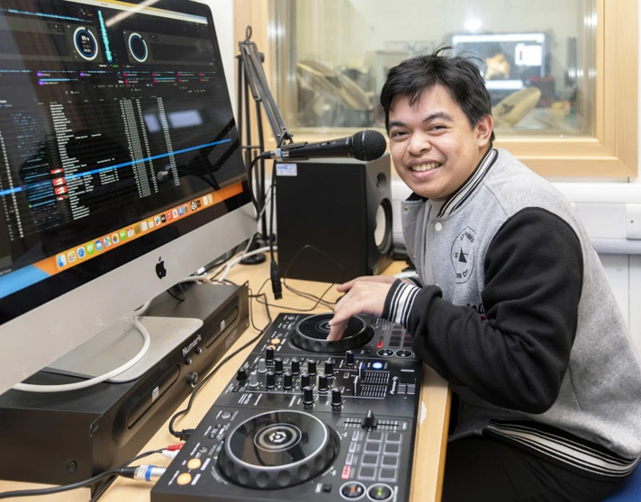 Young man smiling while using DJ equipment and a microphone, with a computer screen displaying music software. Young man smiling while using DJ equipment and a microphone, with a computer screen displaying music software.